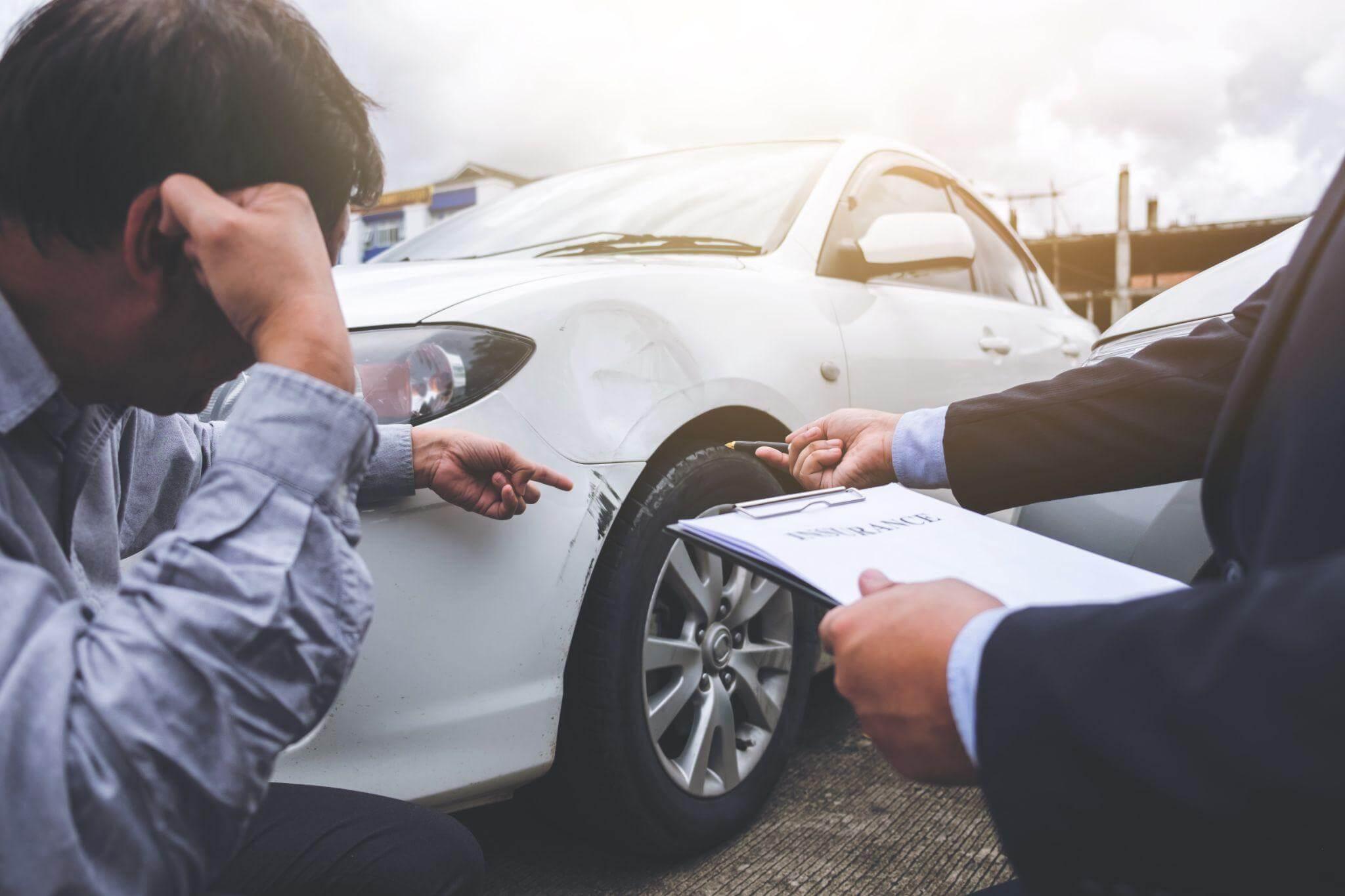 a man sitting next to a white car talking to an insurance adjuster after a car crash 